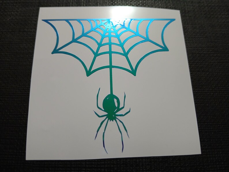 May include: A white sticker featuring a teal and green spider web design with a hanging spider. The spider and web have a gradient effect, transitioning from teal to green and purple. The sticker is square-shaped.