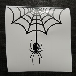 May include: A black spider and web design on a white background. The spider is hanging from the center of the web. The web is a stylized, symmetrical design. The image is a sticker or decal.