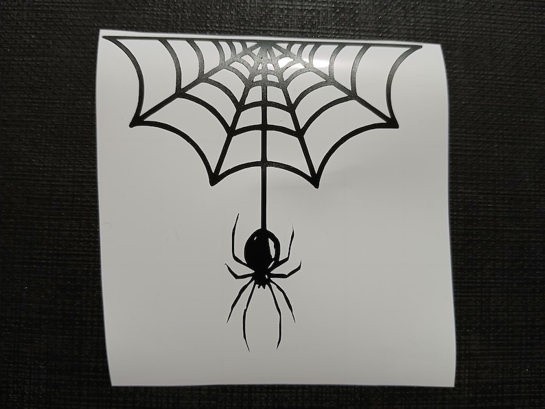 May include: A black spider and web design on a white background. The spider is hanging from the center of the web. The web is a stylized, symmetrical design. The image is a sticker or decal.