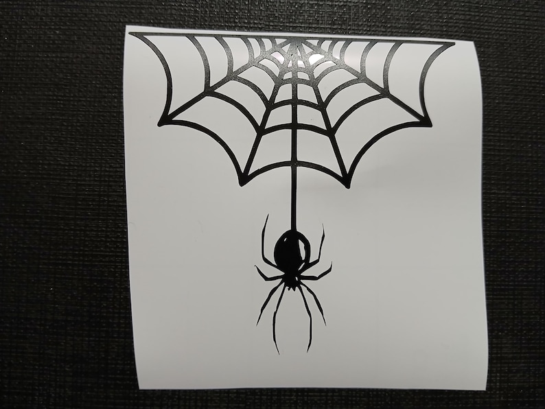 May include: A black spider and web design on a white background. The spider is hanging from the web, which is a detailed, symmetrical pattern. The image is a sticker or decal, perfect for Halloween decorations.