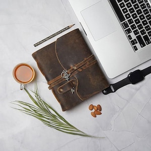May include: A brown leather journal with a key charm and a strap closure. The journal is on a white surface with a laptop, a pencil, a cup of tea, almonds, and a watch.