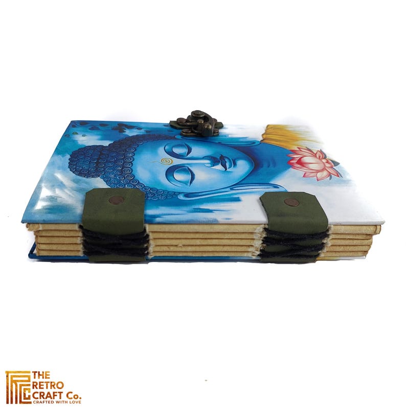 May include: A blue and white journal with a Buddha image on the cover. The journal has a green leather strap and a metal clasp. The journal is handmade and has a rustic look.