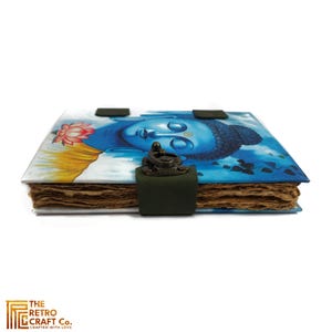 May include: A blue and green journal with a painting of Buddha on the cover. The journal has a leather strap closure and a metal lock.