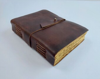 May include: A brown leather-bound journal with a strap closure and stitching detail. The journal is closed and lying on a white surface.