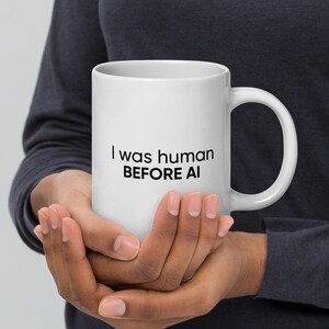 Op de afbeelding: Witte keramische mok met de tekst "I was human BEFORE AI" in het zwart. De mok heeft een groot oor en wordt door een persoon vastgehouden.