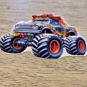 May include: A sticker of a monster truck decorated with colorful Christmas lights and a red bow on top. The truck has large black and white tires with orange rims.