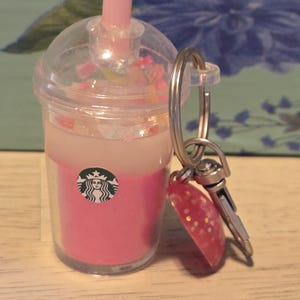 May include: A miniature Starbucks-style drink keychain with a pink and white layered liquid, a pink straw, and a clear lid. The keychain includes a red charm with gold glitter and a silver key ring.