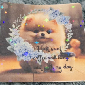 May include: A square sticker featuring a Pomeranian dog surrounded by blue flowers and greenery. The text "I just want coffee and hang out with my dog" is printed on the sticker. The sticker has a holographic finish.