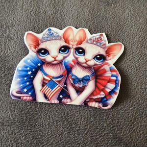 May include: A sticker featuring two cartoon Sphynx cats adorned with patriotic attire. One cat holds a miniature American flag. They wear jeweled crowns and are dressed in red, white, and blue outfits, celebrating a national holiday.