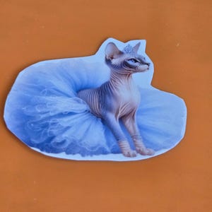 May include: A sticker featuring a Sphynx cat wearing a blue tutu and a tiara. The cat is hairless with blue eyes and is sitting with its front paws forward. The background is a light blue color.