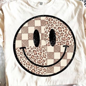 May include: Cream-colored t-shirt featuring a large smiley face design. The smiley face is split into two halves: one with a brown and cream checkered pattern and the other with a leopard print. The smiley face has black eyes and a black smile.