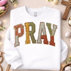 Op de afbeelding: Wit sweatshirt met het woord "PRAY" in patchwork letters. De letters zijn in verschillende patronen en kleuren, waaronder paisley, ruitjes en bloemenmotieven. Paasdecoraties omringen het sweatshirt.