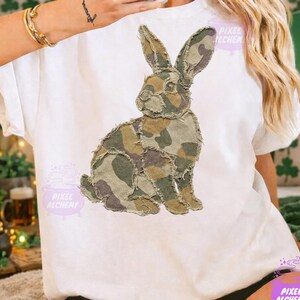 May include: White t-shirt featuring a large, appliqued rabbit design in camouflage fabric. The rabbit is detailed with shades of green, brown, and tan. The shirt has short sleeves and a crew neck. The brand name "Pixel Alchemy" is visible.