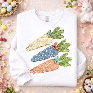 May include: White sweatshirt featuring three carrot designs. Each carrot has a unique pattern: yellow with daisies, blue with white polka dots, and orange with swirls. Green leaves and bows adorn the carrots, perfect for Easter.