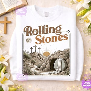 May include: White sweatshirt featuring a graphic with the text "Rolling Stones" in brown, with a scene of three crosses on a hill, a rising sun, and a tomb. The design includes a full moon and a stone. The sweatshirt has a crew neck.