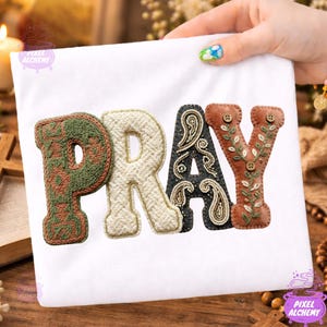 Op de afbeelding: Wit T-shirt met het woord "PRAY" geborduurd in getextureerde, kleurrijke letters. De letters zijn in tinten groen, crème, zwart en bruin, met decoratieve patronen en knoopaccenten. Het shirt wordt vastgehouden door een hand met gelakte nagels.