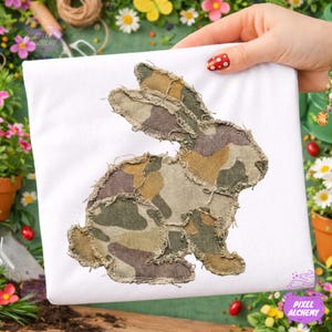 May include: White t-shirt with a camouflage bunny design. The bunny is made from patches of green, brown, and tan fabric with frayed edges. The t-shirt is held up by a hand with red polka dot nail polish. The Pixel Alchemy logo is visible.