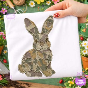 May include: A white t-shirt featuring a detailed applique of a rabbit. The rabbit is crafted from camouflage fabric, with visible pocket details, and button eyes. The design is centered on the shirt, showcasing a unique, handcrafted aesthetic.