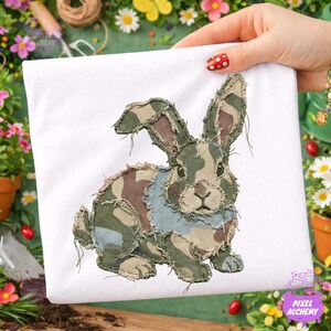 May include: White t-shirt with a patchwork rabbit design in camouflage shades of green, brown, and blue. The rabbit has long ears and is centered on the shirt. The design has a distressed, textured appearance.