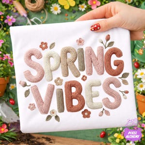 May include: White t-shirt with the words "SPRING VIBES" in a knitted style, featuring various pastel colors. The design includes small floral and leaf accents, creating a spring-themed aesthetic.