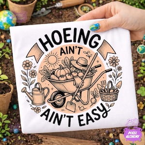 以下が含まれることがあります： 円形のデザインが施された白いTシャツ。野菜でいっぱいの手押し車、じょうろ、そして「HOEING AIN'T AIN'T EASY」というテキストが描かれています。デザインには太陽、花、園芸道具が含まれています。