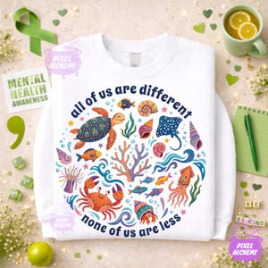 May include: White sweatshirt with a colorful sea life design. The graphic includes a turtle, fish, coral, and other sea creatures, with the text "all of us are different, none of us are less". The sweatshirt promotes mental health awareness.