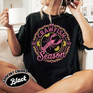 May include: Black t-shirt with a pink and yellow graphic that says "Crawfish Season." The graphic features a pink crawfish and lemon slices. The shirt also has a "Comfort Colors Black" label.