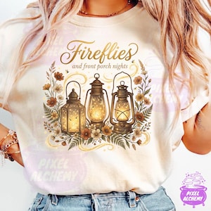 May include: Cream-colored t-shirt featuring a graphic of three vintage lanterns with glowing lights, surrounded by flowers and foliage. The text "Fireflies and front porch nights" is written above the lanterns in a decorative font.