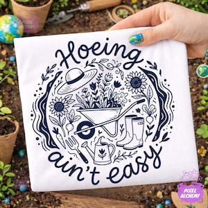 May include: White t-shirt with a navy blue gardening-themed graphic. The design includes a wheelbarrow, sunflowers, a watering can, and the text "Hoeing ain't easy". The design is surrounded by a circular border of leaves and flowers.