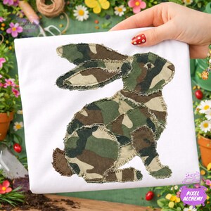 May include: White t-shirt featuring a seated rabbit applique made of camouflage fabric. The rabbit has long ears and a fluffy tail. The design is stitched onto the shirt with a frayed edge.