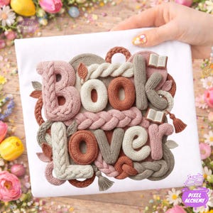 May include: White t-shirt featuring the text "BOOK LOVER" in a 3D knitted style. The letters are in shades of pink, brown, and grey, with small open book details. The design is surrounded by decorative knitted elements.