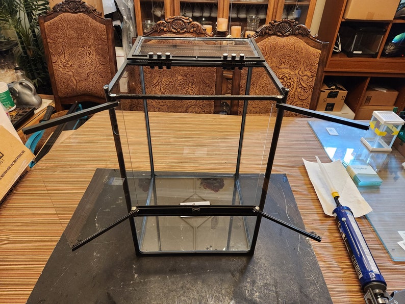 May include: A black glass terrarium with a hinged top and removable sides. The terrarium is made of clear glass and has a black metal frame. The terrarium is sitting on a wooden table.