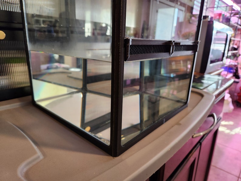 May include: A black framed glass terrarium with a black mesh screen on the top. The terrarium is filled with water and is sitting on a gray surface.