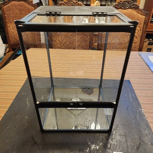 May include: A black glass terrarium with two shelves and a hinged lid. The terrarium is empty and sits on a wooden surface.