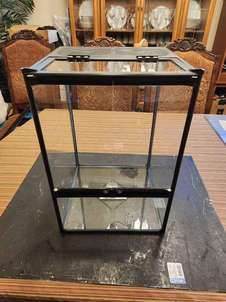May include: A black glass terrarium with two shelves and a hinged lid. The terrarium is empty and sits on a wooden surface.