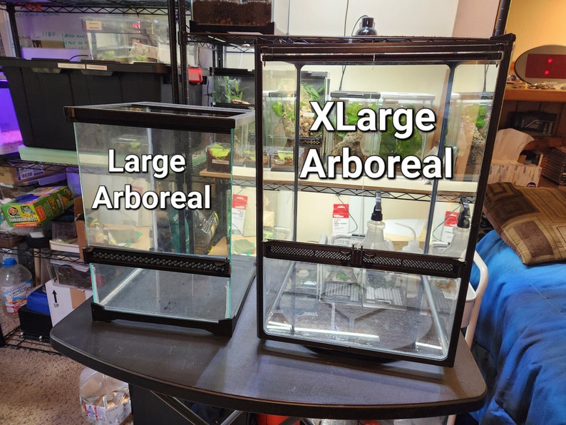 May include: Two black glass terrariums with black metal frames. The smaller terrarium is labeled "Large Arboreal" and the larger terrarium is labeled "XLarge Arboreal".