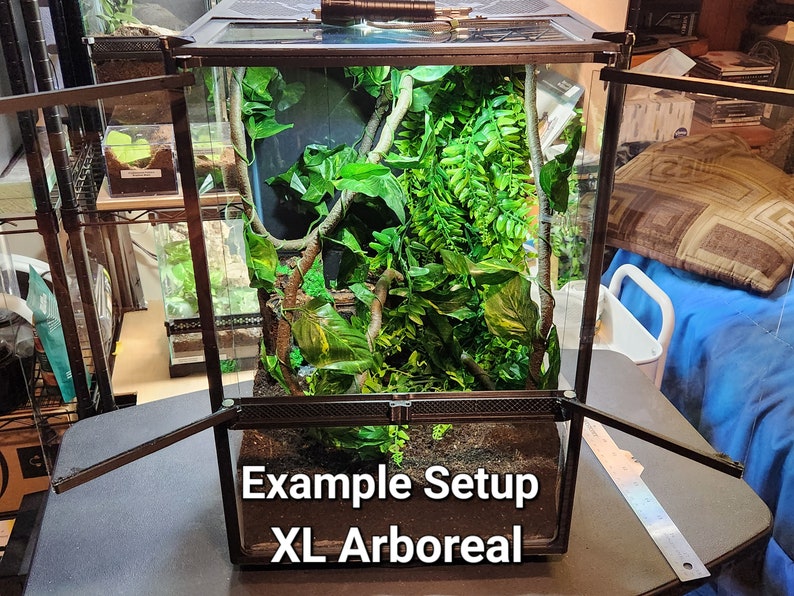 May include: A black glass terrarium with a black metal frame and a black mesh top. The terrarium is filled with artificial plants and vines. The terrarium is labeled "Example Setup XL Arboreal".