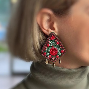 May include: Embroidered diamond-shaped earrings with a green border and a red floral design. The earrings have small gold-colored charms hanging from the bottom. The earrings are worn on the ear.