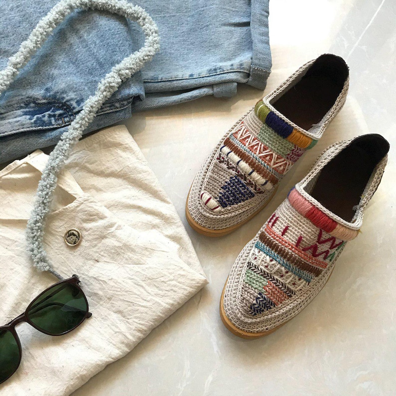 May include: A pair of beige crochet slip-on shoes with colorful embroidered patterns. The shoes have a rounded toe and a flat sole.