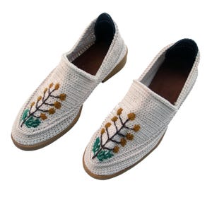 May include: A pair of white crocheted slip-on shoes with embroidered floral designs in green and brown.