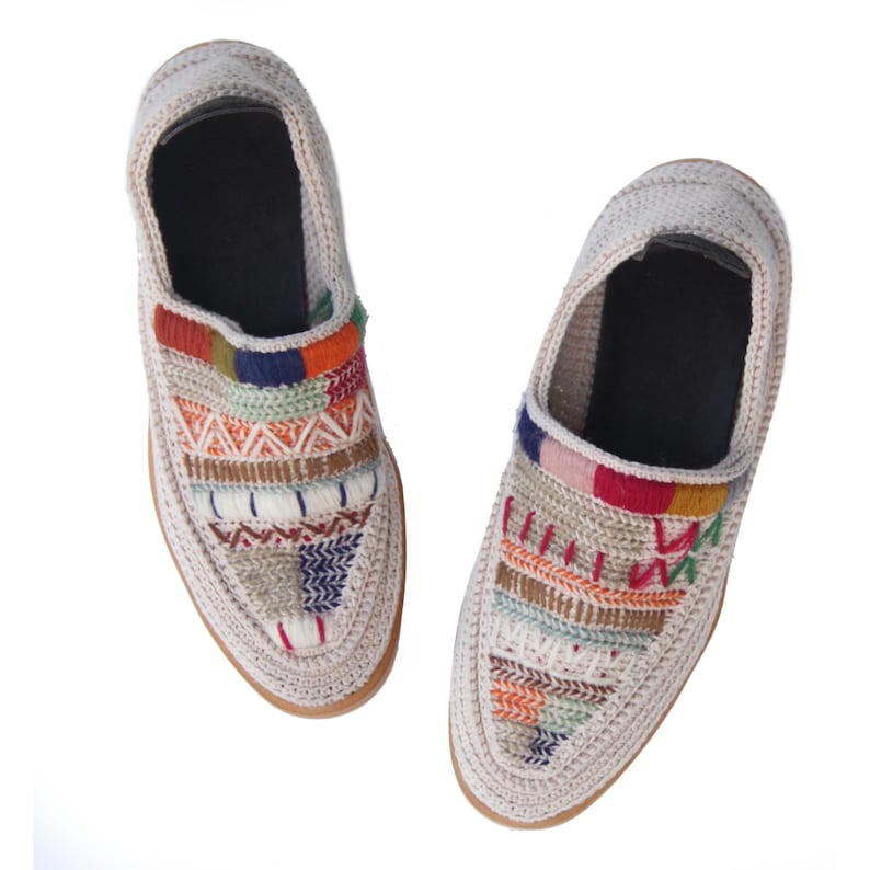 May include: A pair of beige crocheted slip-on shoes with colorful embroidered patterns. The shoes have a flat sole and a rounded toe.