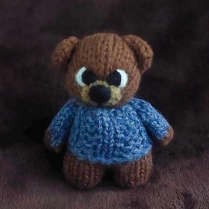 May include: A handmade brown teddy bear wearing a blue knitted sweater. The bear has white eyes, a black nose, and a tan muzzle. The bear is standing on a brown surface.