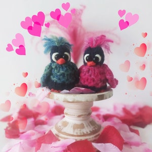 May include: Two small crocheted birds, one blue and green, the other pink, are perched on a white pedestal cake stand. The stand is surrounded by pink flower petals.