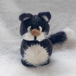Könnte beinhalten: Eine handgefertigte Miniatur-Katzenfigur. Die Katze ist aus dunkelgrauem und weißem Garn gefertigt, mit einer flauschigen weißen Brust und einem Schwanz. Sie hat kleine, spitze Ohren und eine braune Nase. Die Katze ist auf einer weichen, weißen Oberfläche platziert.