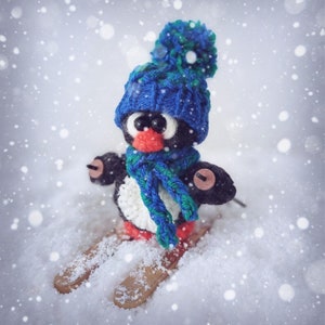 May include: A crocheted penguin wearing a blue knit hat and scarf, skiing down a snowy hill. The penguin has black and white fur and a red beak.