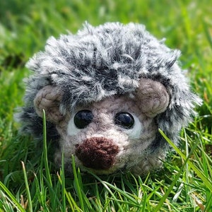 Può includere: Un giocattolo di peluche riccio grigio e marrone con occhi neri e un naso marrone, seduto sull'erba verde.