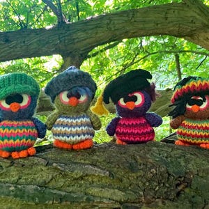 Peut inclure: Quatre figurines de hiboux crochetés à la main, avec des corps rayés colorés, des pieds orange et différents styles de chapeaux. Les hiboux sont perchés sur une branche d'arbre sur fond de feuillage vert. Chaque hibou a de grands yeux ronds.