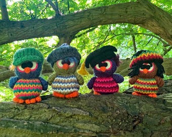 May include: Four handmade crochet owl figurines with colorful striped bodies, orange feet, and various hat styles. The owls are perched on a tree branch against a backdrop of green foliage. Each owl has large, round eyes.