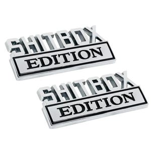 May include: Two silver metal car emblems with black lettering. The emblems read "Shitbox Edition".