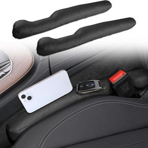May include: Two black car seat gap fillers are shown, along with a white smartphone, car key, and seat belt buckle. The gap fillers are designed to prevent items from falling into the car seat crevices. The interior of a car is visible.